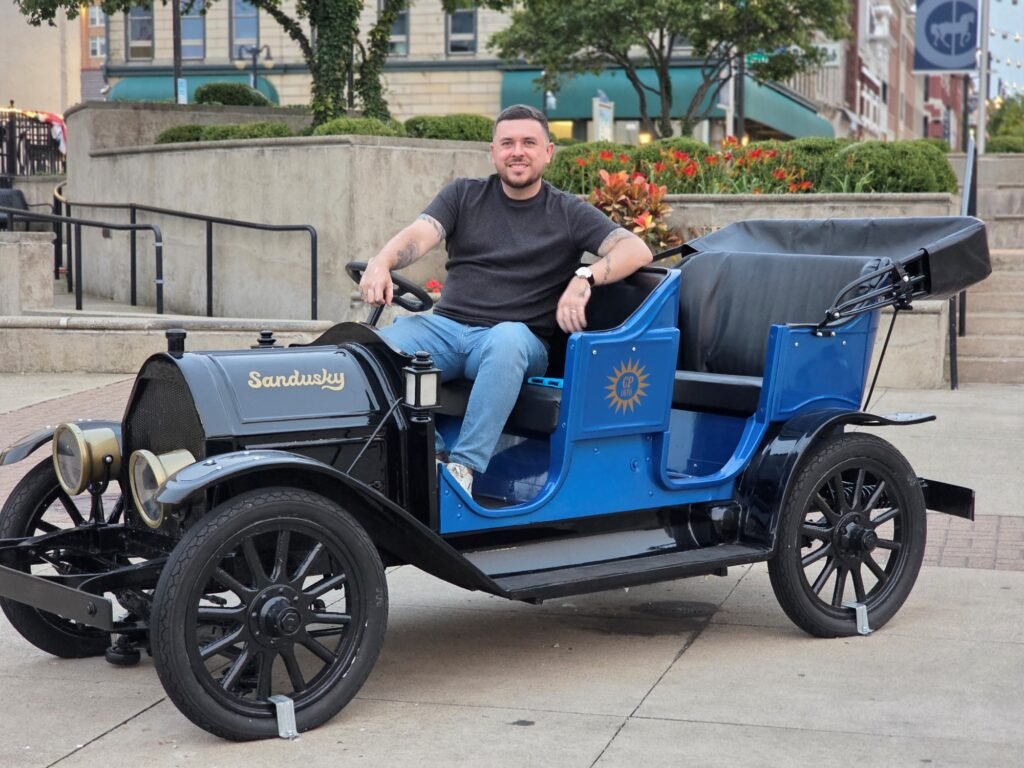 A person sits in a vintage black and blue car with the word "Sandusky" displayed on the front.